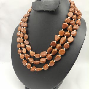 Puede incluir: Un collar de tres hilos con cuentas de piedra de oro de color cobre, pulidas e irregulares, con un aspecto brillante y centelleante. Los hilos están separados por pequeñas cuentas blancas redondas, creando un diseño en capas con un tono metálico cálido.
