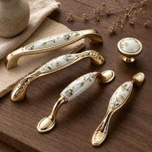 Könnte beinhalten: Eine Sammlung goldfarbener Schrankbeschläge, darunter Griffe und ein Knauf. Die weißen Keramikmittelstücke sind mit einem floralen Muster in Lila und Grün verziert. Die Hardware wird auf einer Holzoberfläche präsentiert.
