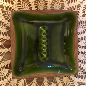 May include: A square-shaped ceramic ashtray featuring a glossy, deep forest green glazed center basin with a central oval indentation containing five small circular cigarette rests, surrounded by a textured, matte brown wood-grain border.