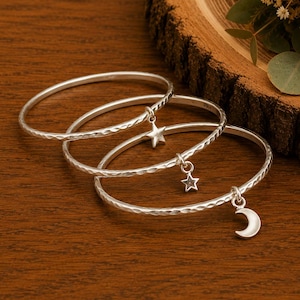 Peut inclure: Trois bracelets joncs argentés avec une texture martelée. Chaque bracelet comporte une petite breloque suspendue : l'un a une étoile à cinq branches, un autre une étoile plus petite, et le troisième une forme de croissant de lune.
