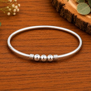 Puede incluir: Una pulsera rígida de tono plateado con una banda circular lisa. El centro de la pulsera está adornado con tres esferas plateadas pulidas, flanqueadas a cada lado por dos pequeños anillos plateados estriados que actúan como espaciadores.