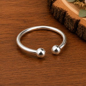 May include: A silver-colored cuff bracelet with a rounded design and two ball-shaped ends. The bracelet is displayed on a wooden surface, with a floral arrangement in the background. The jewelry is a simple, elegant accessory.