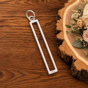 May include: A silver pendant featuring a long, rectangular open frame design with a circular bail at the top. The bottom edge of the rectangular frame is clearly stamped with the hallmark 925, indicating the sterling silver composition of the piece.