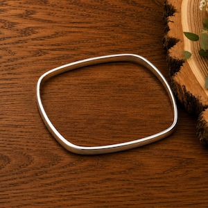 May include: A smooth, polished silver-toned bangle bracelet with a rounded square shape. The bangle features a continuous, seamless metal band with a reflective surface, presented as a single piece of jewelry without any clasps or additional embellishments.