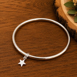 Peut inclure: Un bracelet jonc en argent poli doté d'une unique petite breloque en forme d'étoile à cinq branches fixée à l'anneau circulaire. La surface métallique est lisse et réfléchissante, présentant un design simple et continu avec un pendentif étoile.