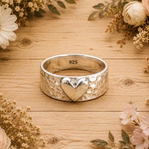 May include: A wide silver ring with a hammered texture and a heart-shaped design. The ring is stamped with "925" and is surrounded by flowers and greenery on a wooden surface.