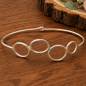 May include: A polished silver-tone bangle bracelet featuring a series of four interconnected circular and oval loops of varying sizes along the front, with a simple hook-and-eye clasp mechanism visible at the back of the thin, rounded metal band.