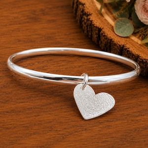 Puede incluir: Una pulsera rígida de plata pulida con un dije en forma de corazón. El dije tiene una superficie texturizada que recuerda a una huella dactilar. La pulsera es un aro circular liso y continuo hecho de un material metálico de tono plateado.