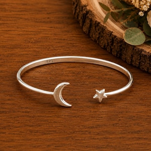 Puede incluir: Una pulsera abierta de plata con un dije de media luna en un extremo y un dije de estrella de cinco puntas en el otro. La banda de metal está estampada con el número 925, lo que indica una composición de plata esterlina, y tiene un acabado liso.
