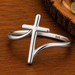 May include: A polished silver-tone ring featuring a prominent cross design integrated into the band. The metal surface is smooth and reflective, with the number 925 clearly engraved on the inner side of the band, indicating sterling silver composition.