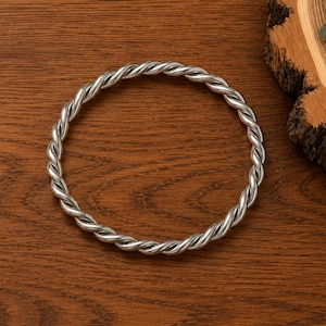Puede incluir: Una pulsera de metal plateado retorcido. La pulsera es circular y se encuentra sobre una superficie de madera marrón oscuro. Un pequeño arreglo floral y vegetación se encuentra sobre una rodaja de madera en el fondo.
