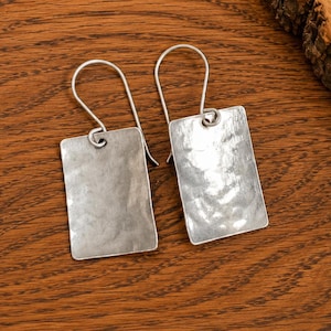 May include: A pair of silver-toned earrings featuring rectangular pendants with a hammered, textured surface. Each pendant is attached to a simple, curved wire hook for suspension, showcasing a metallic finish with subtle light reflections.