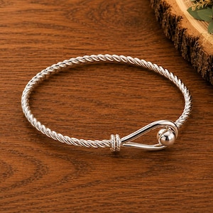 Puede incluir: Una pulsera de tono plateado con una banda de estilo cuerda trenzada que forma un bucle en un extremo. El cierre consiste en una cuenta esférica lisa y pulida que asegura el bucle, creando un diseño circular continuo con acabado metálico.