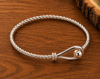 May include: A silver-toned bracelet featuring a twisted rope-style band that forms a loop at one end. The closure consists of a smooth, polished spherical bead that secures the loop, creating a continuous circular design with a metallic finish.