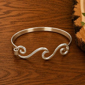 May include: A polished silver bangle bracelet featuring a continuous wave design with three distinct crests. The metal has a smooth, reflective finish and a circular shape. The bracelet is constructed from a single piece of silver-toned metal.