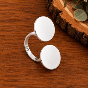 May include: A silver open ring featuring two smooth, flat circular discs at each end of the band. The inner side of the band is clearly stamped with the hallmark 925, indicating the sterling silver composition of this polished jewellery piece.