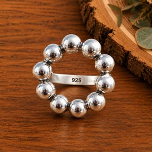 May include: A polished silver ring featuring a circular arrangement of ten smooth, spherical beads. The band of the ring is clearly stamped with the hallmark 925, indicating the sterling silver composition of this reflective, metallic jewelry piece.
