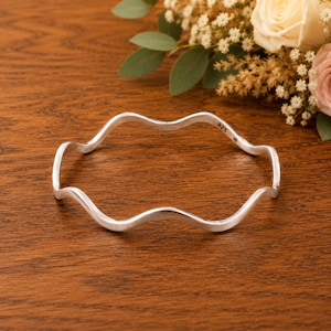 May include: A silver bangle bracelet with a continuous wavy, undulating design. The inner surface of the metal band features a clear stamp reading 925, indicating sterling silver composition, along with a small, distinct maker's mark symbol.