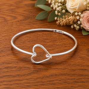 Könnte beinhalten: Ein polierter Silberarmreif mit einem mittigen, offenen Herzdesign. Das Metallband ist glatt und kreisförmig und weist auf der Innenseite einen kleinen Stempel mit einem Blumensymbol und der Zahl 925 auf, was auf Sterlingsilber hinweist.