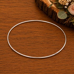 May include: A thin, circular silver bangle bracelet with a smooth, polished finish. The metal band features a small, clear engraving of the number 925, indicating its sterling silver composition, visible along the lower right curve of the piece.