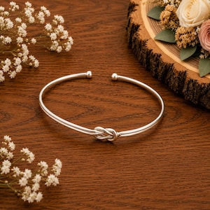 May include: A silver-toned open cuff bracelet featuring a central knot design and small spherical terminals at each end. The bracelet has a smooth, polished metallic finish and a circular shape, presented as a single piece of jewelry.