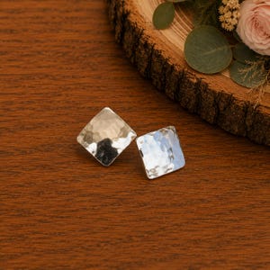 May include: A pair of square-shaped stud earrings with a polished, hammered silver finish. Each earring features a distinct textured surface with multiple indentations that reflect light, creating a bright, metallic appearance on the flat metal.