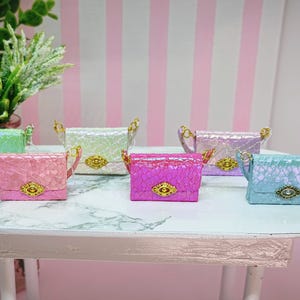 Puede incluir: Una colección de pequeños bolsos de mano coloridos en tonos rosa, verde y azul. Cada bolso presenta una superficie texturizada, herrajes dorados y un asa pequeña. Los bolsos están colocados sobre una mesa blanca. Los bolsos miden unos 5 cm.