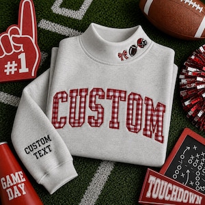May include: A light gray mock neck sweatshirt featuring the word CUSTOM in red and white gingham applique letters across the chest. The collar includes small embroidered football, bow, and helmet icons. The sleeve displays the text CUSTOM TEXT.