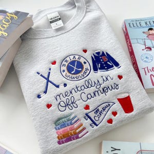 May include: A light gray sweatshirt with embroidered collegiate-themed graphics. The design includes hockey sticks, a varsity jacket, a red cup, books, and the words "Briar University" and "mentally in Off-Campus". Several books are visible in the background.