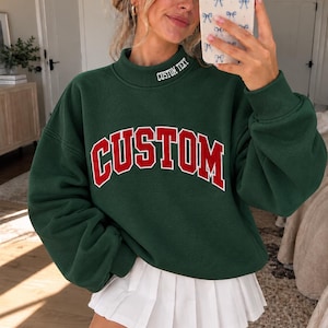 May include: A dark green long-sleeve sweatshirt with a mock neck collar featuring the white embroidered text CUSTOM TEXT. The front of the sweatshirt displays the word CUSTOM in large red block letters with a white outline. A white pleated skirt.
