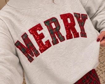 May include: A light gray long-sleeve sweatshirt with a mock neck collar. The word MERRY is appliqued across the chest in large block letters featuring a red and green plaid pattern with red sequin accents on the edges of each letter.