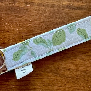 May include: A light blue key fob with a green leaf pattern. The key fob has a silver metal ring and a brown leather accent. A small tag reads "handmade with".