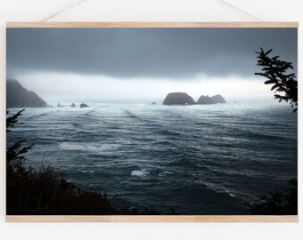 May include: A moody seascape photograph featuring a dark, stormy sky with a few clouds and a distant view of rocky islands. The ocean is a deep blue with whitecaps and waves. The image is framed with a light wood frame.