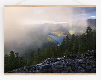 May include: A misty mountain landscape with a lake in the distance. The sun shines through the clouds, creating a rainbow effect. The foreground is covered in rocks.