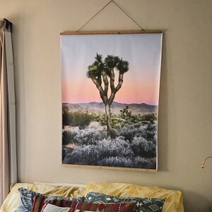 May include: A white bed with a white crocheted bedspread and a variety of patterned throw pillows. A large framed print of a Joshua tree in a desert landscape hangs on the wall above the bed.