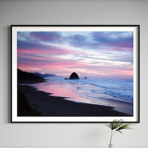 Puede incluir: Una fotografía enmarcada de una escena de playa con una gran formación rocosa en la distancia. El cielo es de un rosa y un morado vibrantes, reflejándose en el agua. La playa es arenosa y las olas rompen en la orilla.