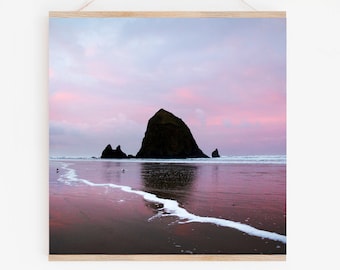 May include: A silhouette of Haystack Rock, a large sea stack, on a beach at sunset. The sky is a vibrant pink and purple, reflecting in the calm water.