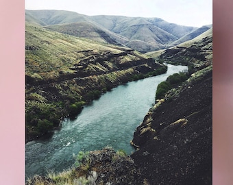 May include: A framed print of a landscape photograph featuring a river winding through a valley. The river is a vibrant blue-green color, and the valley is a mix of green and brown hues. The print is mounted on a wooden frame.