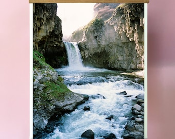 May include: A wooden framed poster with a photograph of a waterfall cascading down a rocky cliffside. The water flows into a river with rapids and large rocks. The poster is hanging from a black cord.