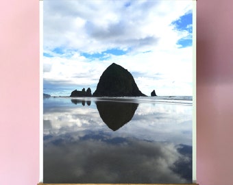 Puede incluir: Una impresión en lienzo enmarcada en madera de una gran formación rocosa en el océano con un reflejo de la roca y las nubes en el agua. El cielo es azul con nubes blancas.