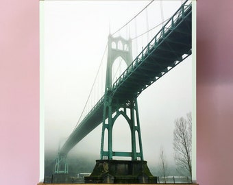 May include: A framed print of a green bridge in a foggy setting. The bridge is a suspension bridge with a large arch in the center. The print is hanging from a wooden frame.