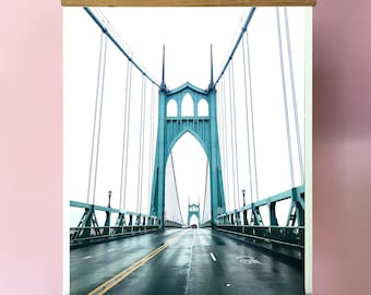 May include: A canvas print of the Steel Bridge in Portland, Oregon. The bridge is a green steel structure with a road running through the center. The print is framed with a wooden frame and has a rope hanger.
