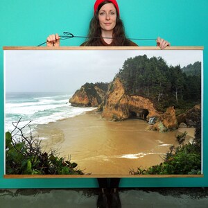 May include: A panoramic photograph of a rocky coastline with a sandy beach and crashing waves. The photo is framed in a wooden frame and is being held by a person wearing a red hat.