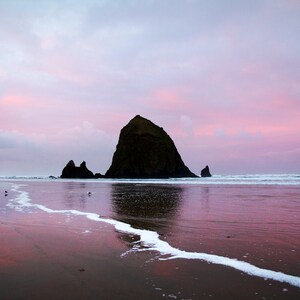 May include: A large rock formation, known as Haystack Rock, stands tall against a pink and purple sunset sky. The rock is reflected in the calm, still water of the ocean. The beach is sandy and wet, with white foam from the waves.