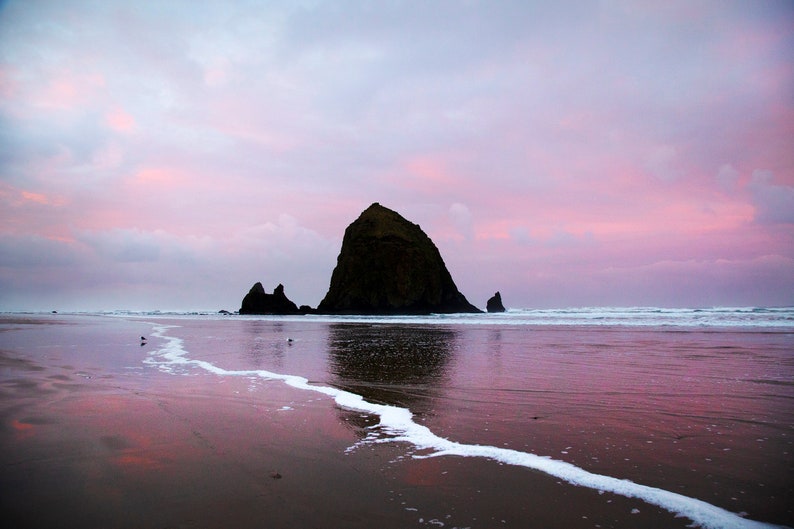 May include: A large rock formation, known as Haystack Rock, stands tall against a pink and purple sunset sky. The rock is reflected in the calm, still water of the ocean. The beach is sandy and wet, with white foam from the waves.