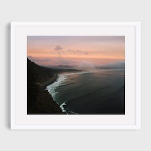 May include: A panoramic view of a rocky coastline with a misty sky and a vast ocean in the distance. The sky is a soft pink and purple, with wispy clouds. The ocean is a deep blue, with white waves crashing on the shore. The rocky coastline is a dark brown, with green vegetation growing on the slopes.