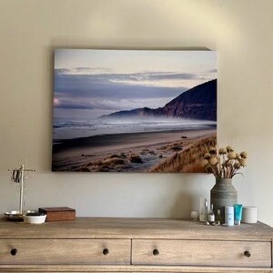 May include: A framed photograph of a beach scene with a misty, cloudy sky and a mountain range in the background. The beach is sandy with waves crashing on the shore.