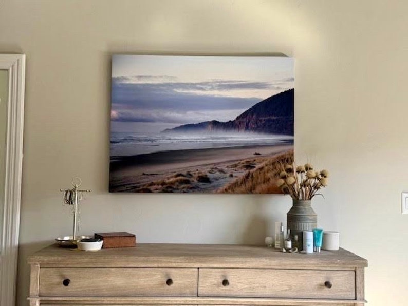May include: A framed photograph of a beach scene with a misty, cloudy sky and a mountain range in the background. The beach is sandy with waves crashing on the shore.