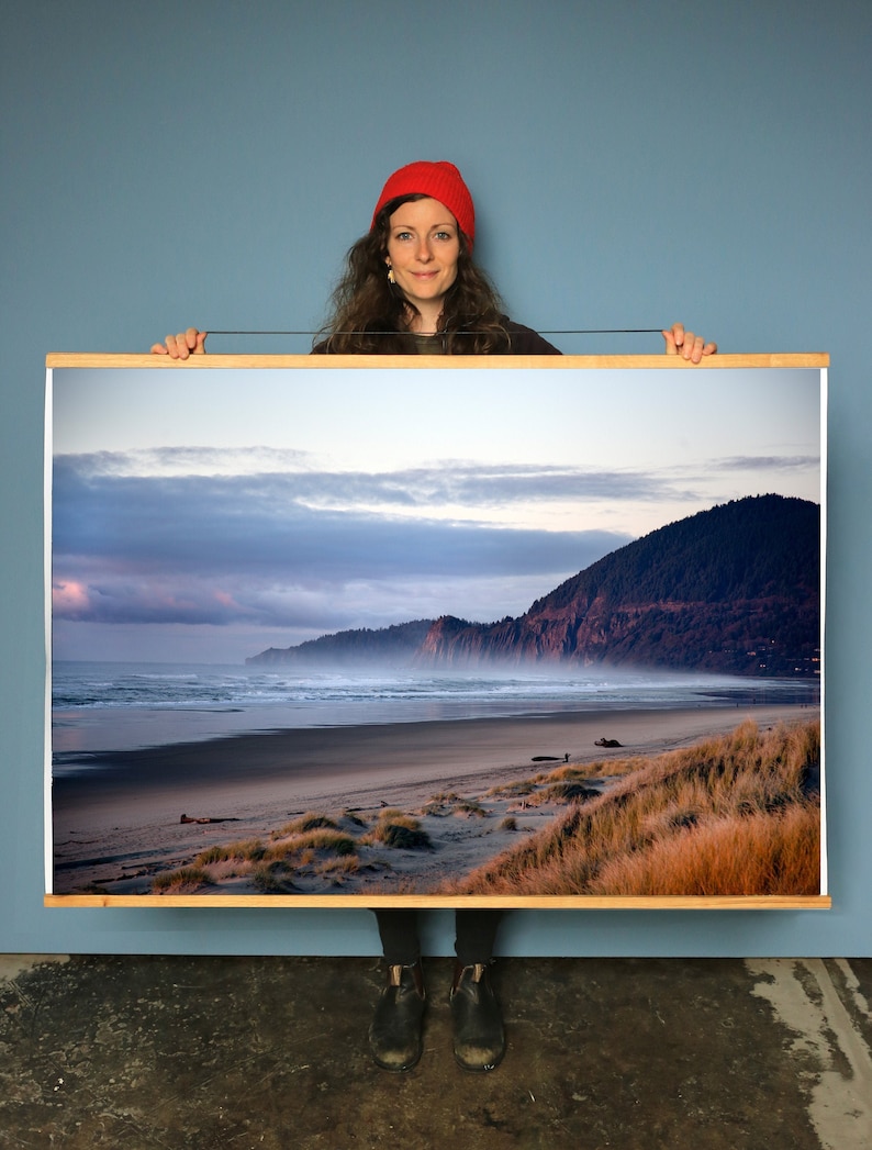 May include: A framed photograph of a beach scene with a rocky cliff in the distance. The sky is a mix of blue, gray, and pink, and the sand is a light brown. The photo is hanging on a wall, and a person is holding it up.