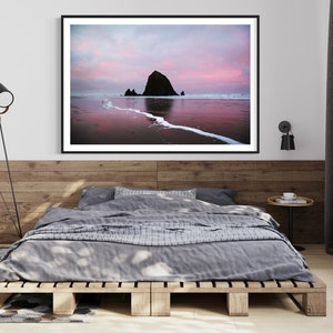 May include: A framed photograph of a large rock formation on a beach at sunset. The sky is a vibrant pink and orange, and the water is reflecting the colors of the sky. The rock formation is silhouetted against the sky.